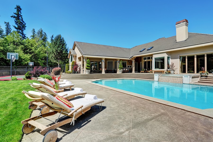 View of a luxury home with a pool on a sunny day with outdoor lounge chairs and a basketball court in the background.