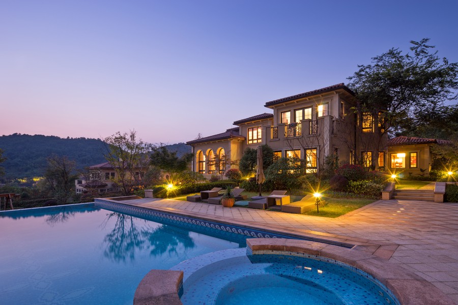 A well-lit home at dusk with a pool in the foreground and small hill in the background. 