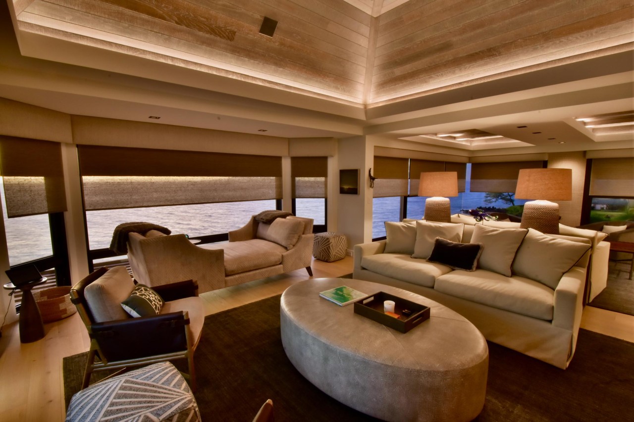 Modern living room with floor-to-ceiling windows showcasing motorized shades, comfortable beige seating, and warm layered lighting creating a cozy ambiance by the water.