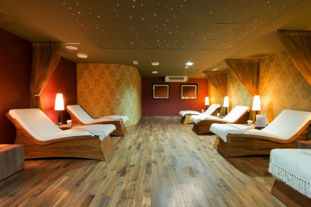 A tranquil spa relaxation room featuring wooden wave-shaped loungers with white cushions. The room has warm lighting from small lamps, gold patterned wallpaper, and a ceiling designed with tiny sparkling lights to mimic a starry night.