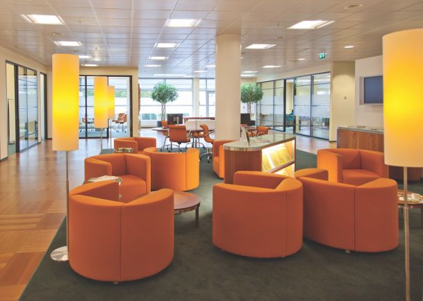 A modern open-plan office with a cluster of orange tub chairs on a dark green rug. Three tall, glowing floor lamps provide warm lighting, and a white support pillar stands in the center. In the background, there are glass-walled meeting rooms, potted indoor trees, and light wood flooring under a grid of recessed ceiling lights. 